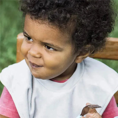 A child with curly hair is wearing a light-colored bib, holding a fork and smiling while sitting outdoors.