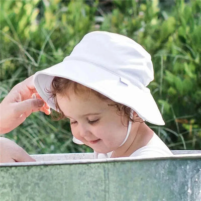 Baby wearing a white breathable bucket hat with chin strap, smiling and interacting with an adult hand in a natural setting.