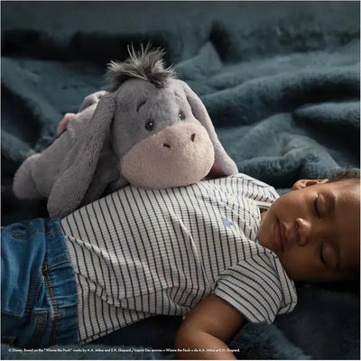 Gund Oh So Snuggly Eeyore plush toy rests on a sleeping child wearing a striped shirt, surrounded by soft blankets.