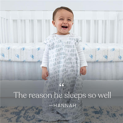 Baby wearing a HALO SleepSack Micro-Fleece blanket with a gray pattern, smiling next to a crib with whale-patterned bedding.