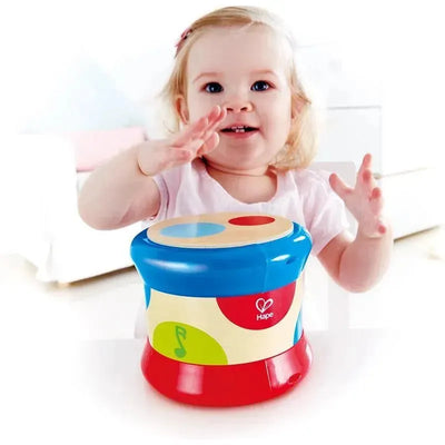 Toddler happily plays with a colorful Hape baby drum, reaching out with hands, in a bright, playful environment.
