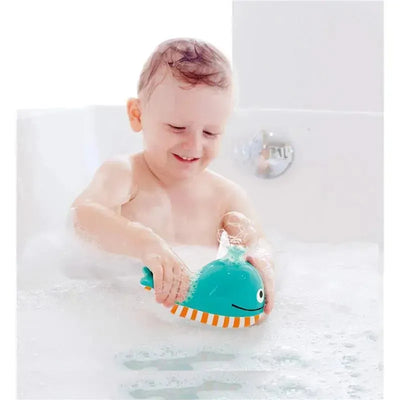 A child in a bubble-filled bath joyfully plays with the Hape Bubble Blowing Whale, a teal whale toy with orange stripes.
