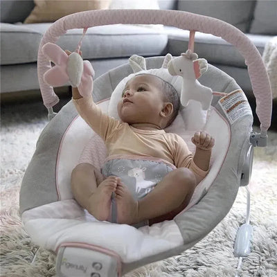 Baby in a soothing bouncer seat reaching for hanging toys, featuring a plush unicorn and flower, with a soft, padded design.