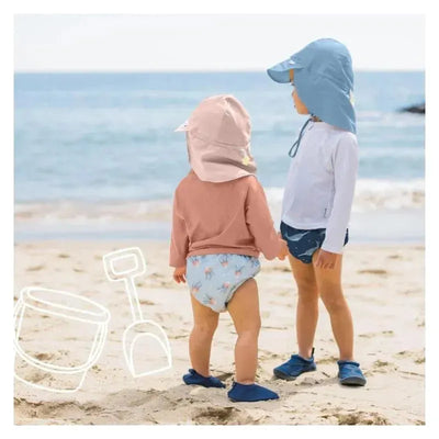 Two children play on the beach, one wearing a light lavender swim diaper with a floral pattern, the other in a blue outfit.