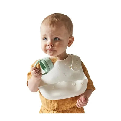 A baby in a sage green feeder holds a silicone Snacker while wearing a bib, looking curiously to the side.