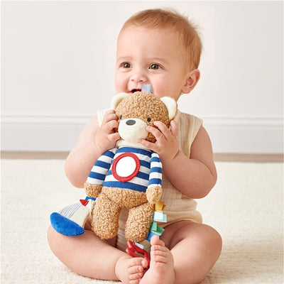 Baby joyfully holds the Itzy Ritzy Bear sensory toy, featuring a striped shirt and colorful ribbons for tactile exploration.
