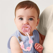 Baby with a pink bow silicone pacifier and clip, wearing a blue outfit, held by an adult.
