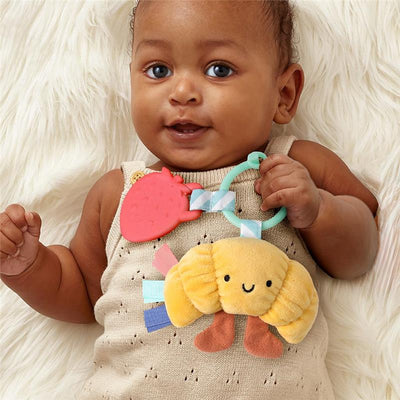 Infant holding a colorful Itzy Ritzy teething toy with a plush croissant and a strawberry-shaped teether.