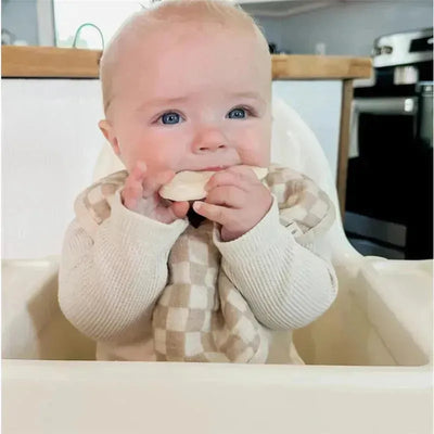 Baby sitting in a high chair wearing a beige checkered bib, chewing on a teething accessory, with a cozy kitchen backdrop.