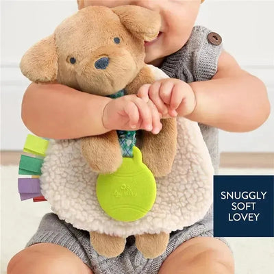 Plush Puppy Itzy Friends Lovey™ being held by a smiling baby, featuring colorful tags and a green teething attachment.