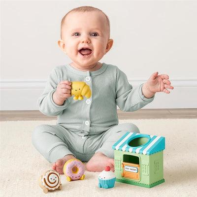 Smiling baby in a green romper holding a yellow toy near a colorful bakery playset with various food figures.
