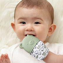 Baby smiling while holding an Itzy Ritzy Sage Bear Teething Mitt, featuring a green bear design with a fabric sleeve.