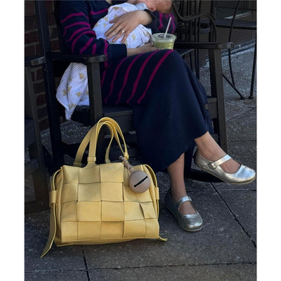 Yellow woven handbag resting on the ground next to a seated woman holding a wrapped baby, with a beverage nearby.