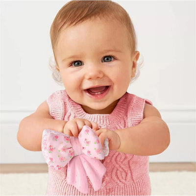 Smiling baby holding a pink silicone sensory teether shaped like a bow with floral patterns, wearing a light pink sweater.