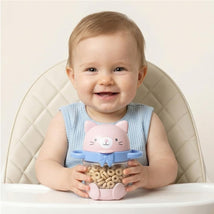Baby sitting in a high chair, happily holding a pink kitty-themed snack cup filled with cereal rings.