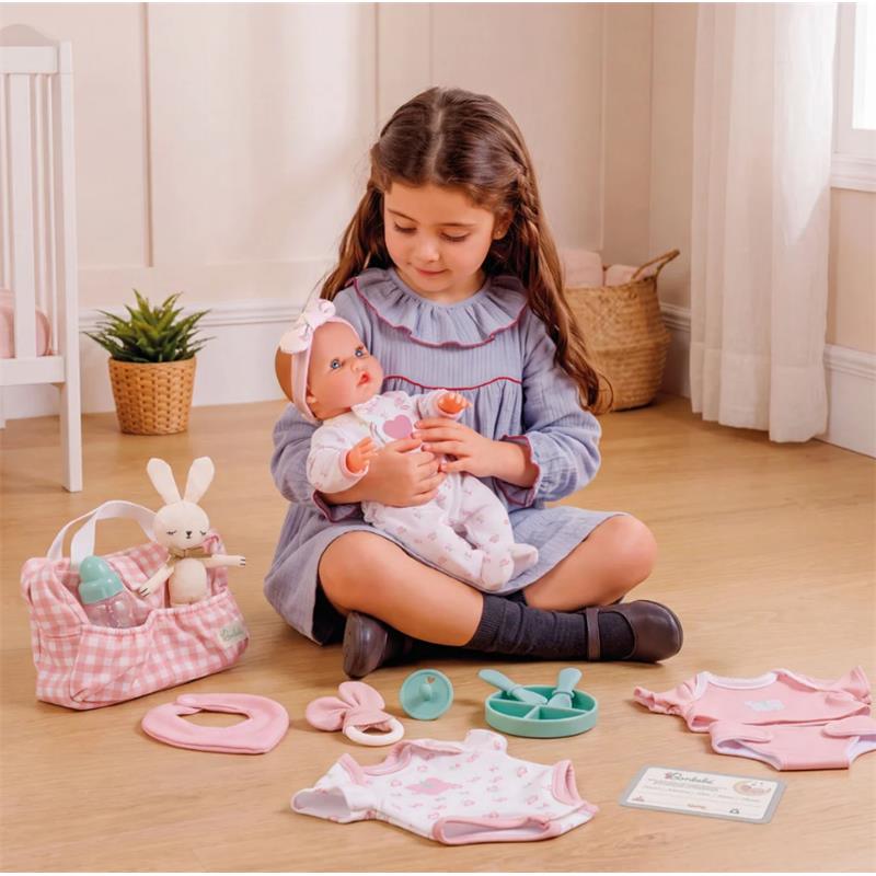 A girl sitting on the floor holds a 15.5” Bonbebé weighted reborn baby doll among various play accessories.