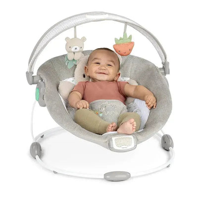 Baby sitting in the Ingenuity InLighten Baby Bouncer Seat, smiling, with toys hanging above and a soft gray seat.