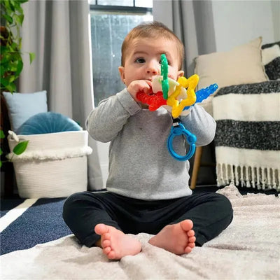 Toddler sitting on a rug, exploring a colorful Sea Of Sensory Teether Toy with a blue attachment clip.
