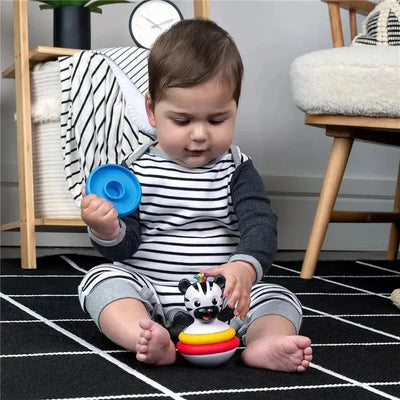 Baby sitting on a black and white rug, playing with the Kids II Stack & Wobble Zen Teether Toy, a zebra-shaped stacking toy with colorful rings.