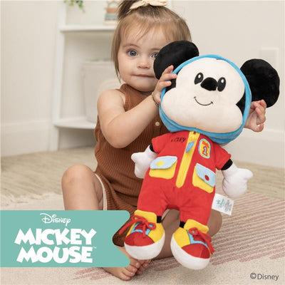 A toddler holds a plush Mickey Mouse toy, wearing a red outfit with blue accents, on a light carpeted surface.