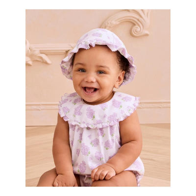 Smiling baby wearing a lilac floppy hat and a matching sleeveless outfit with floral patterns, sitting on a wooden floor.