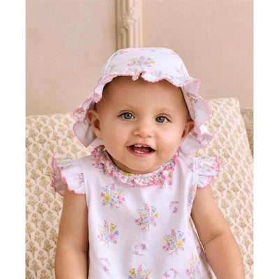 Smiling baby wearing a Kissy Kissy Pastel Gardens floppy hat with floral patterns, sitting on a patterned sofa.