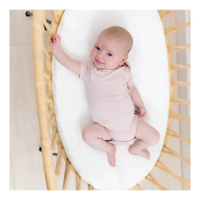 Baby wearing a soft blush Kyte Baby bodysuit, smiling while lying on a cozy white mattress in a woven bassinet.