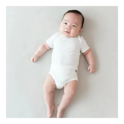 Neutral white baby bodysuit worn by an infant lying on a light surface, smiling with visible arms and legs.
