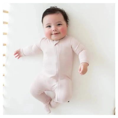 Baby in a blush zippered footie, lying on a light surface, smiling with arms stretched out.