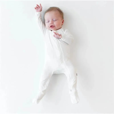Baby lying on a white surface, wearing a white zippered footie. The baby looks relaxed with arms raised.