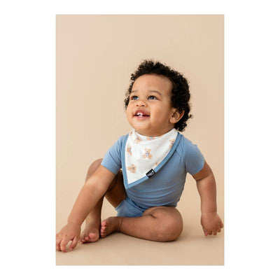 Toddler wearing a blue onesie and a white bib featuring teddy bear designs, smiling while sitting on a beige background.