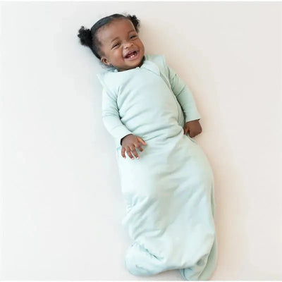 Baby smiling while lying down in a soft sage green Kyte Baby bamboo sleep bag, designed for comfort.