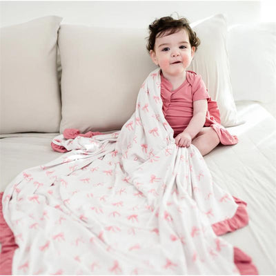 Toddler sitting on a bed, holding a Kyte Baby Ruffle Toddler Blanket featuring a bow pattern in pink and white.