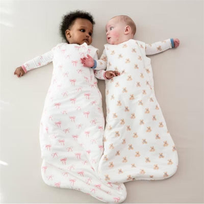 Two babies in Kyte Baby Sleep Bags: one with a pink bow design, the other with bear prints, lying side by side on a light surface.
