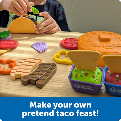 A child assembles a taco using colorful play food, with various ingredients like lettuce, tomatoes, and taco shells on a wooden table.
