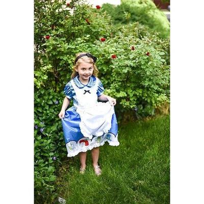 A young girl in a blue Alice in Wonderland dress holds the skirt, smiling in a garden with greenery and red flowers.