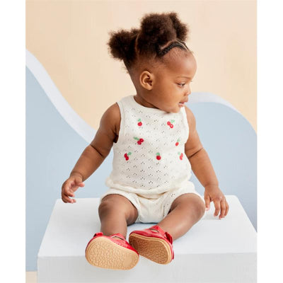 Baby girl in an ivory cherry pointelle knit set, seated on a pedestal, wearing red shoes, with a soft background.