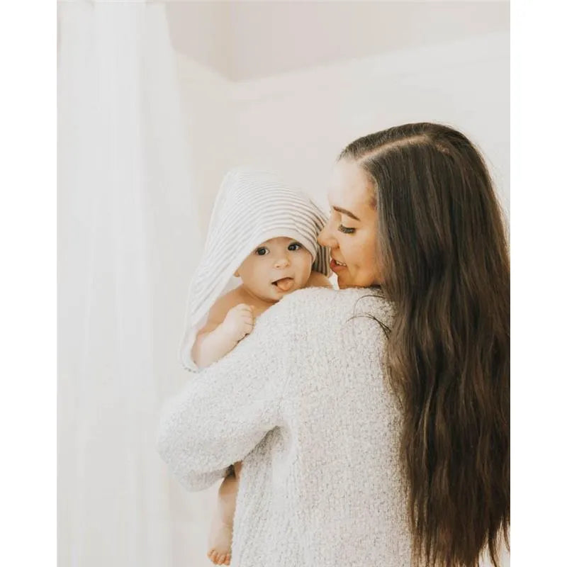 A mother holds her baby, who is wrapped in a grey striped hooded towel, both smiling warmly while surrounded by soft, light-colored surroundings.