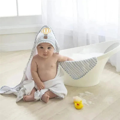 Baby wearing a hooded towel with a hot air balloon design, sitting on a bathroom floor next to a bathtub and rubber duck.