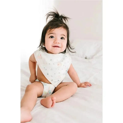 Infant seated on a bed wearing a floral muslin bib and patterned diaper, smiling at the camera.
