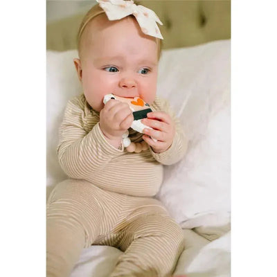 A baby in beige striped pajamas bites a colorful Ebi Shrimp teether while sitting on a bed.