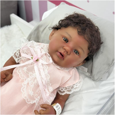 Reborn Baby Doll Shyann, an African American vinyl doll wearing a pink dress with lace and a bow, resting on a grey pillow.