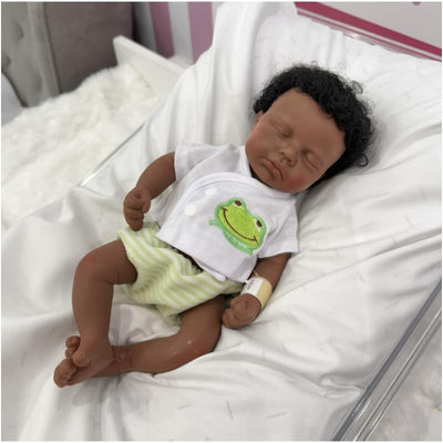 Full silicone 13-inch African American reborn baby doll with rooted hair, wearing a frog-themed outfit and lying on a soft blanket.