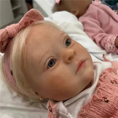 Reborn baby doll with white vinyl, blonde hair, blue eyes, and wearing a pink outfit and headband.