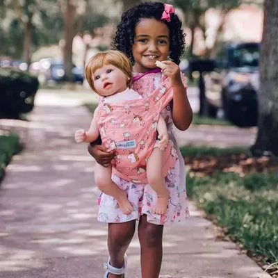 A girl smiles while walking on a path, holding a doll in a pink maternity toy carrier.