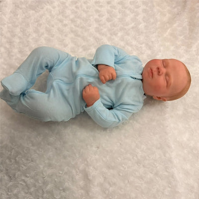 Macrobaby Reborn Doll named Charlie, dressed in a light blue onesie, lying peacefully on a soft white surface.