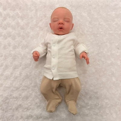 Reborn doll named Megan, made of white vinyl, with painted hair, open blue eyes, dressed in a white long-sleeve onesie and beige pants.