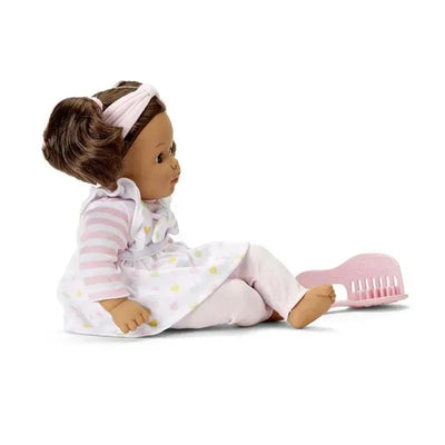 Medium-skinned Madame Alexander doll dressed in a striped shirt and heart-patterned dress, sitting beside a pink comb.