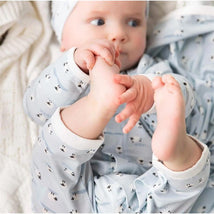 Baby in blue modal coverall with sheep print, playfully holding feet while lounging on a soft blanket.