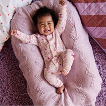 Smiling baby in a pink, patterned convertible coverall, lying on a cozy pink blanket, celebrating with raised arms.